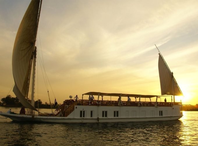 Une grande embarcation blanche de type Dahabiya avec deux voiles triangulaires déployées navigue sur le Nil. Le soleil se couche à l'horizon, créant une lumière dorée intense qui illumine l'eau et le ciel. On aperçoit des silhouettes de passagers sur le pont supérieur en bois