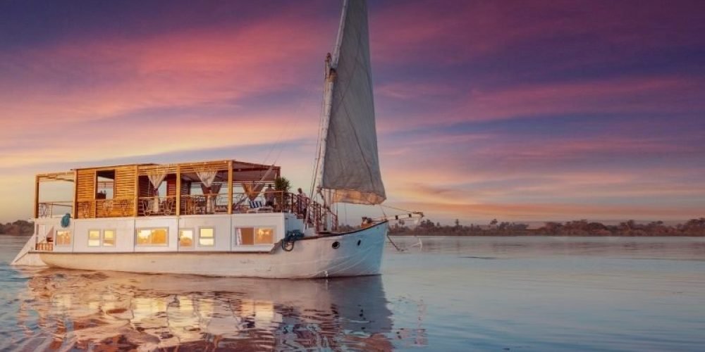 Croisière Dahabiya depuis Assouan 1 Un élégant voilier traditionnel en bois blanc, appelé Dahabiya, navigue sur les eaux calmes du Nil. Le bateau possède une grande voile triangulaire grise à l'avant et une structure de pont avec des rideaux blancs. Le ciel en arrière-plan affiche des nuances magnifiques de violet et d'orange lors d'une Dahabiya depuis Assouan