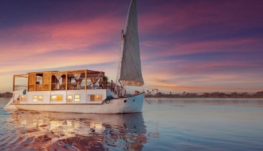Un élégant voilier traditionnel en bois blanc, appelé Dahabiya, navigue sur les eaux calmes du Nil. Le bateau possède une grande voile triangulaire grise à l'avant et une structure de pont avec des rideaux blancs. Le ciel en arrière-plan affiche des nuances magnifiques de violet et d'orange lors d'une Dahabiya depuis Assouan
