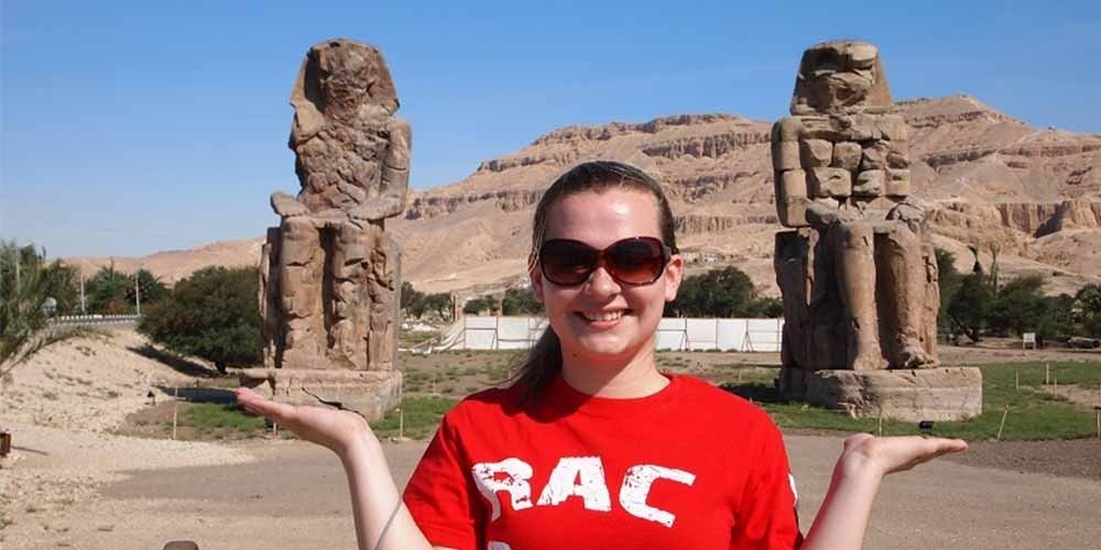 Croisière Dahabiya depuis Assouan 3 Une jeune femme souriante avec des lunettes de soleil et un t-shirt rouge se tient au premier plan, les mains ouvertes. Derrière elle s'élèvent deux statues géantes de pharaons assis sur des trônes de pierre, avec des montagnes désertiques en arrière-plan lors d'une Dahabiya depuis Assouan