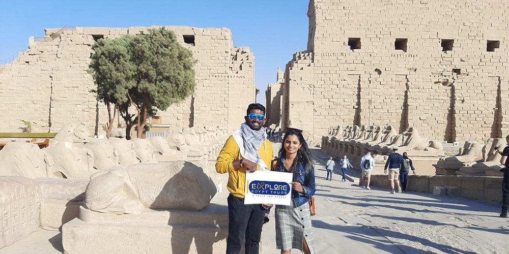 Croisière Dahabiya depuis Assouan 4 Un jeune homme avec un chèche et une femme tenant une pancarte "Explore Egypt Tours" posent devant une allée de sphinx à tête de bélier en pierre. Derrière eux se dressent les murs massifs et les pylônes du temple de Karnak lors d'une Dahabiya depuis Assouan