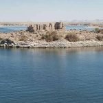 Croisière MS Prince Abbas sur le lac Nasser1 Une vue panoramique prise depuis l'eau montrant une petite île rocheuse au milieu d'une vaste étendue d'eau bleue. Sur l'île، on voit les vestiges en pierre d'un temple antique ou d'une structure historique en ruines، entourés de quelques buissons secs. À l'horizon، on aperçoit des terres désertiques arides sous un ciel clair au-dessus de le lac Nasser