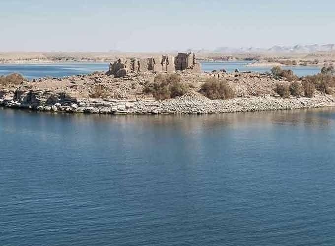 Une vue panoramique prise depuis l'eau montrant une petite île rocheuse au milieu d'une vaste étendue d'eau bleue. Sur l'île، on voit les vestiges en pierre d'un temple antique ou d'une structure historique en ruines، entourés de quelques buissons secs. À l'horizon، on aperçoit des terres désertiques arides sous un ciel clair au-dessus de le lac Nasser