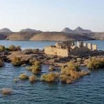 Croisière MS Prince Abbas sur le lac Nasser2 Une vue aérienne rapprochée d'une petite île rocheuse au milieu d'une eau bleue scintillante. L'île abrite les ruines d'un ancien temple égyptien en pierre beige, dont les murs partiels sont encore debout. On voit des arbustes et des buissons secs dispersés sur le sol rocheux, avec des collines désertiques au loin sous un ciel clair au-dessus de le lac Nasser
