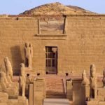 Croisière MS Prince Abbas sur le lac Nasser3 Une vue frontale d'un temple antique en pierre beige avec une entrée centrale protégée par une grille métallique. Devant l'entrée, une allée est bordée de plusieurs statues de faucons sculptées dans la pierre. Sur le mur du temple, on voit des gravures de figures pharaoniques, et une colline rocheuse s'élève juste derrière l'édifice au bord de le lac Nasser