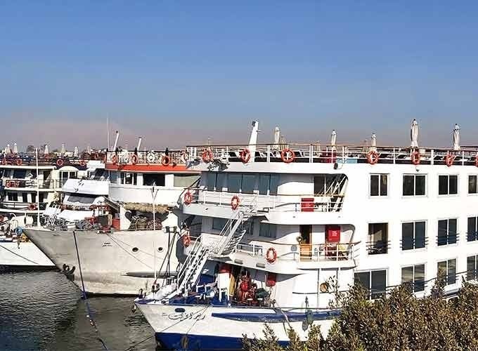Une vue latérale de plusieurs grands navires de croisière blancs amarrés côte à côte sur les eaux calmes du Nil. Les bateaux possèdent plusieurs étages avec des rangées de fenêtres sombres et des ponts supérieurs équipés de garde-corps orange, le tout sous un ciel bleu limpide pour votre Croisière depuis Assouan à Louxor