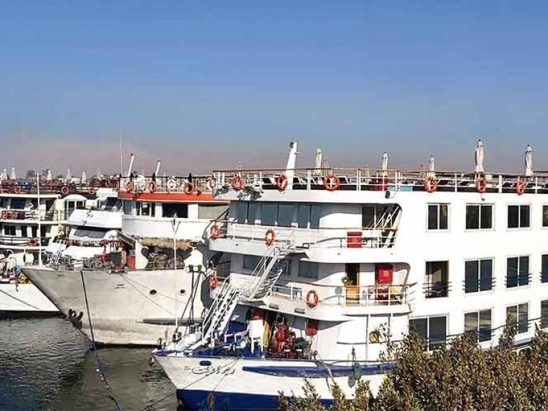 Une vue latérale de plusieurs grands navires de croisière blancs amarrés côte à côte sur les eaux calmes du Nil. Les bateaux possèdent plusieurs étages avec des rangées de fenêtres sombres et des ponts supérieurs équipés de garde-corps orange, le tout sous un ciel bleu limpide pour votre Croisière depuis Assouan à Louxor