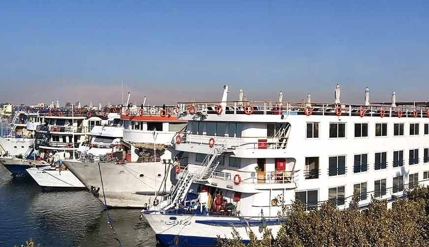 Une vue latérale de plusieurs grands navires de croisière blancs amarrés côte à côte sur les eaux calmes du Nil. Les bateaux possèdent plusieurs étages avec des rangées de fenêtres sombres et des ponts supérieurs équipés de garde-corps orange, le tout sous un ciel bleu limpide pour votre Croisière depuis Assouan à Louxor