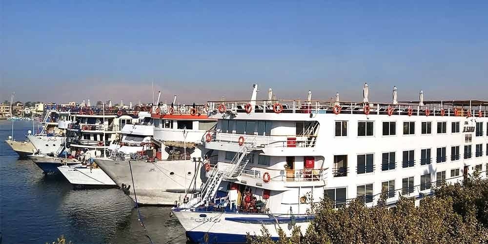 Une vue latérale de plusieurs grands navires de croisière blancs amarrés côte à côte sur les eaux calmes du Nil. Les bateaux possèdent plusieurs étages avec des rangées de fenêtres sombres et des ponts supérieurs équipés de garde-corps orange, le tout sous un ciel bleu limpide pour votre Croisière depuis Assouan à Louxor
