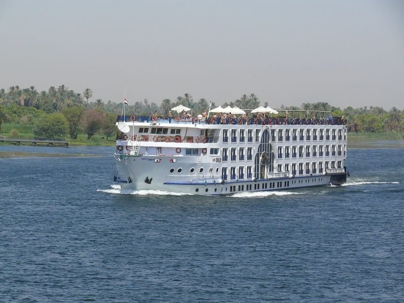 Un grand bateau blanc de plusieurs étages navigue sur les eaux bleues du Nil. Le navire possède de nombreuses fenêtres alignées et un pont supérieur avec des parasols blancs où l'on aperçoit des passagers. À l'arrière-plan, une rive verdoyante avec de nombreux palmiers complète ce décor de Croisière