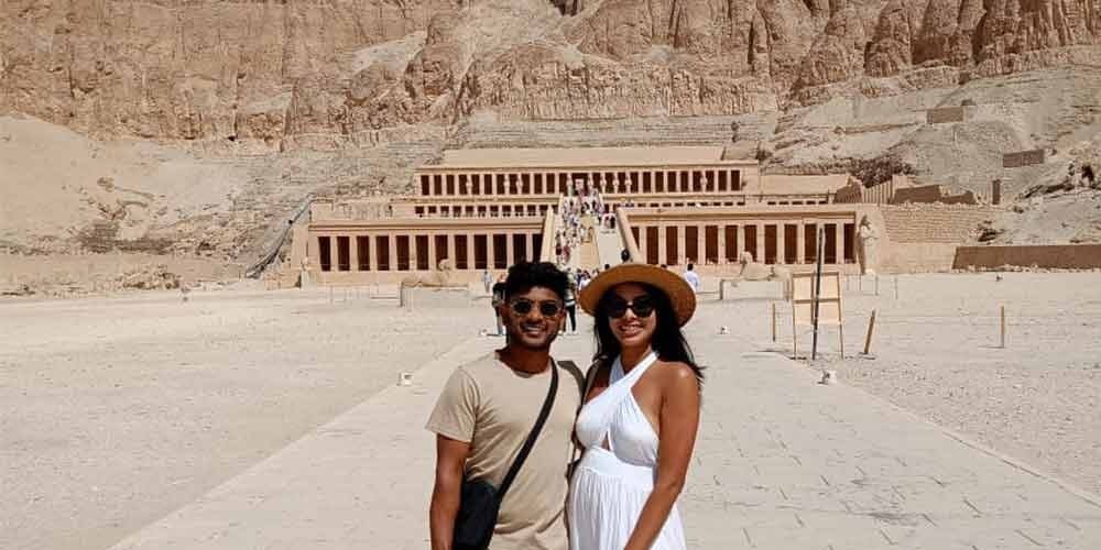 Um casal sorridente posa na rampa de pedra central do Templo de Hatchepsut, em Luxor. O homem veste uma t-shirt amarela, calções brancos e chapéu. A mulher veste uma blusa branca, uma saia azul comprida e um lenço azul na cabeça. Ao fundo, avistam-se as colunas do templo com estátuas de Osíris e os imponentes penhascos de calcário. Um dos pontos altos de uma viagem ao Egito.