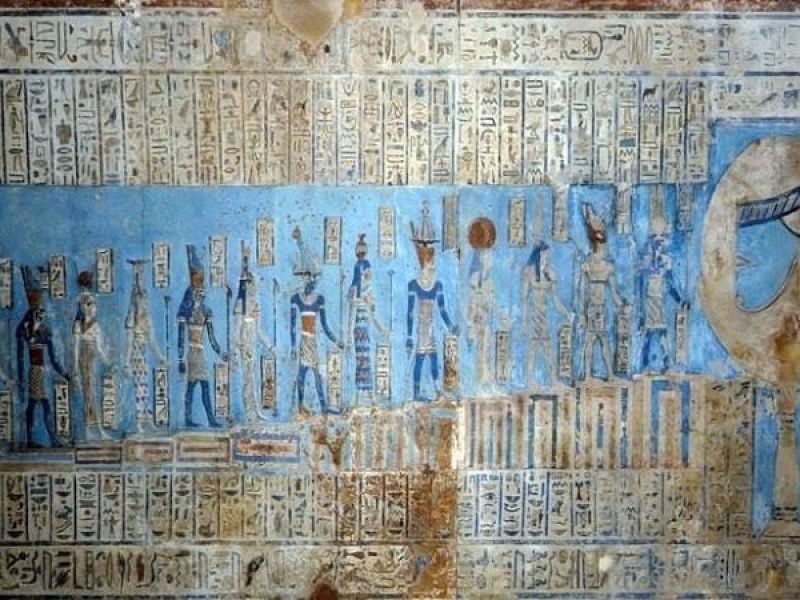 Ancient Egyptian wall relief featuring a procession of deities painted in vibrant blue, surrounded by vertical rows of hieroglyphics and a large Eye of Horus symbol