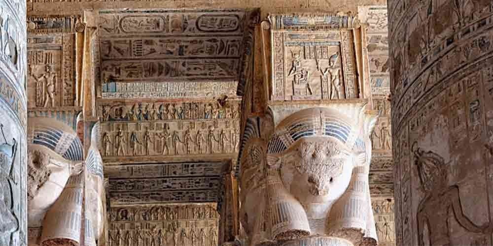 Massive stone columns featuring the carved face of goddess Hathor with cow ears, topped by intricate relief panels showing pharaohs and deities under a decorated temple ceiling