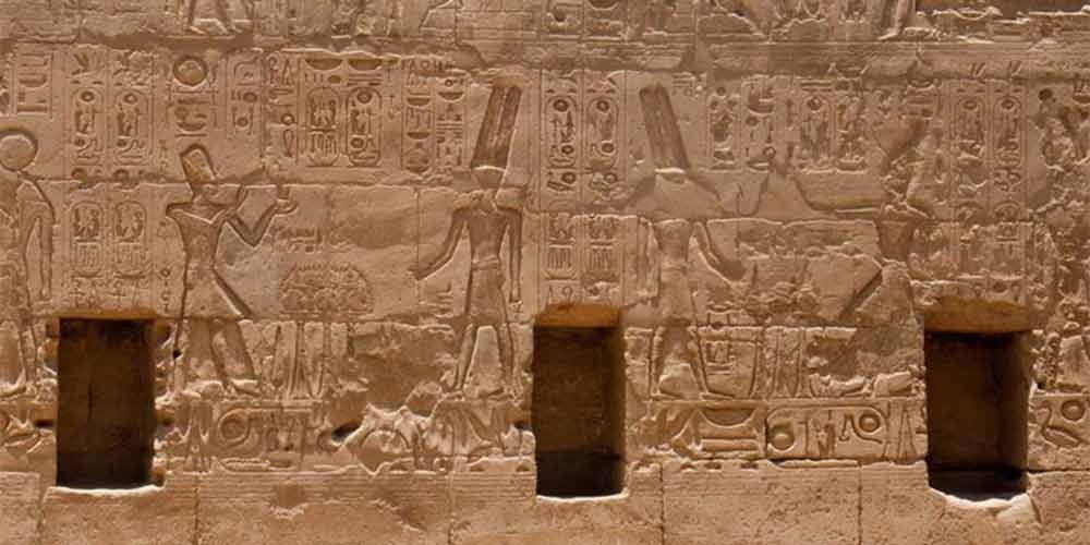 Detailed sandstone wall relief featuring ancient Egyptian figures in traditional headdresses standing between three small rectangular niches, surrounded by vertical columns of hieroglyphics