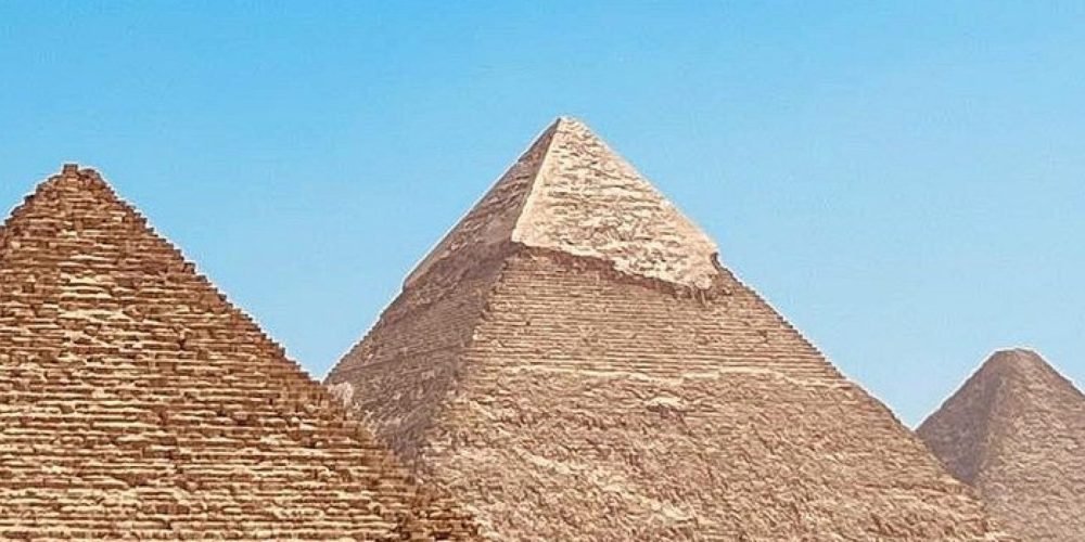 A bright wide-angle shot of the three Great Pyramids of Giza standing majestically in the desert under a clear blue sky. The ancient stone structures show their weathered layers and geometric precision during an Egyptian Museum &Pyramids tour