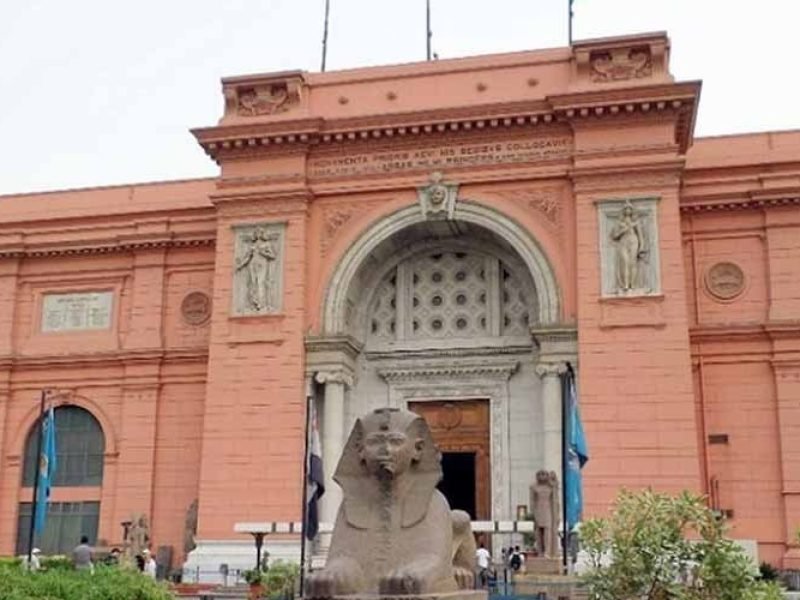 A majestic view of the Egyptian Museum's pink neoclassical facade in Cairo, featuring a grand arched entrance and stone statues. In the foreground, a large stone sphinx sits prominently in front of the historic building during an Egyptian Museum &Pyramids tour