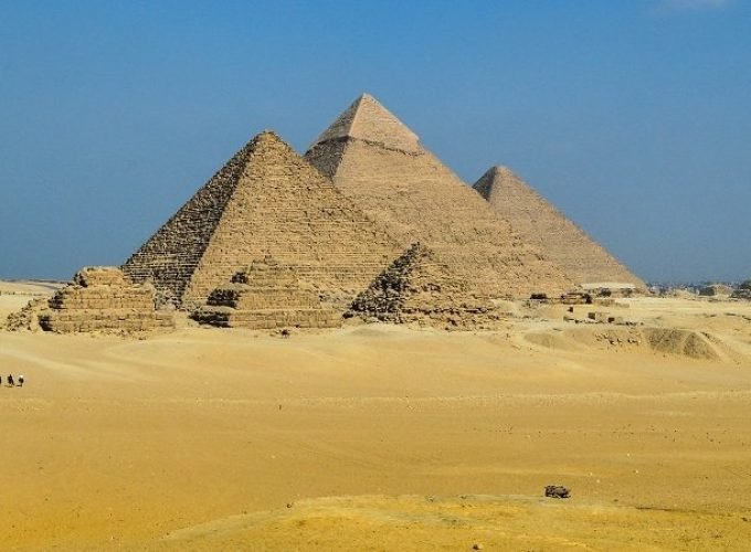A wide landscape of the three Great Pyramids of Giza under a clear blue sky, with several smaller queen pyramids in the foreground. The vast desert sand stretches across the horizon where tourists can be seen walking during their Egyptian Museum trip. The ancient stone structures show detailed textures of the massive blocks used in their historic construction