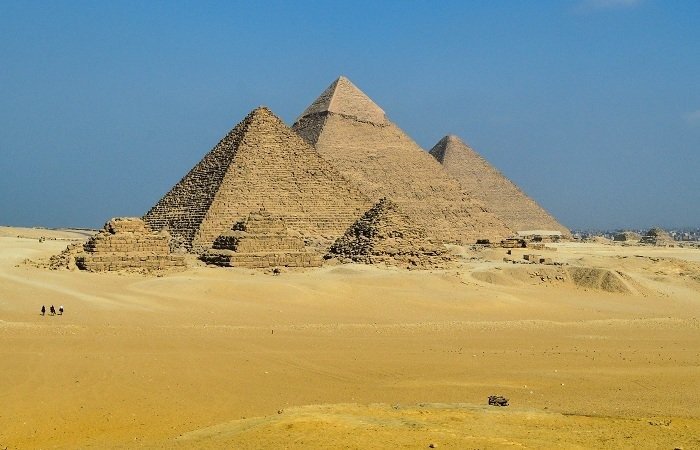 A wide landscape of the three Great Pyramids of Giza under a clear blue sky, with several smaller queen pyramids in the foreground. The vast desert sand stretches across the horizon where tourists can be seen walking during their Egyptian Museum trip. The ancient stone structures show detailed textures of the massive blocks used in their historic construction