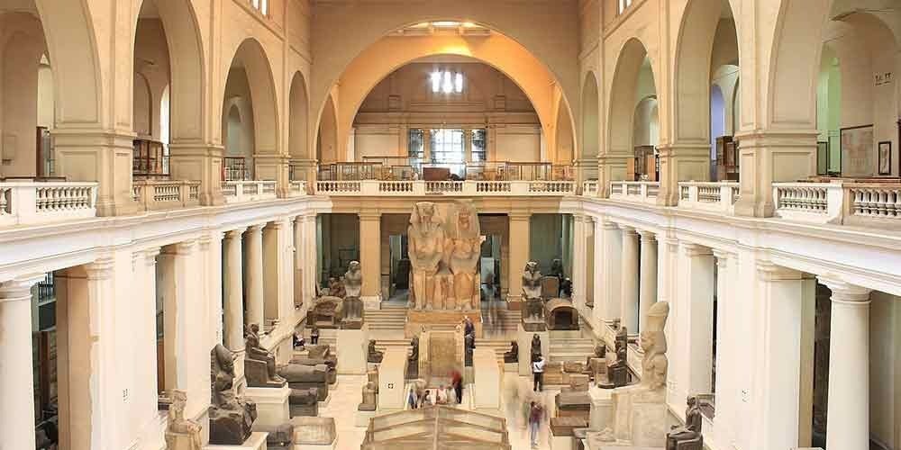 Interior view of the grand hall in the Egyptian Museum featuring high arched ceilings, cream-colored columns, and various ancient Pharaonic statues. Large stone sculptures and seated figures are displayed throughout the floor level of this Egyptian Museum trip, viewed from an upper balcony. Natural light streams into the spacious archaeological gallery, illuminating the diverse collection of artifacts and historic monuments