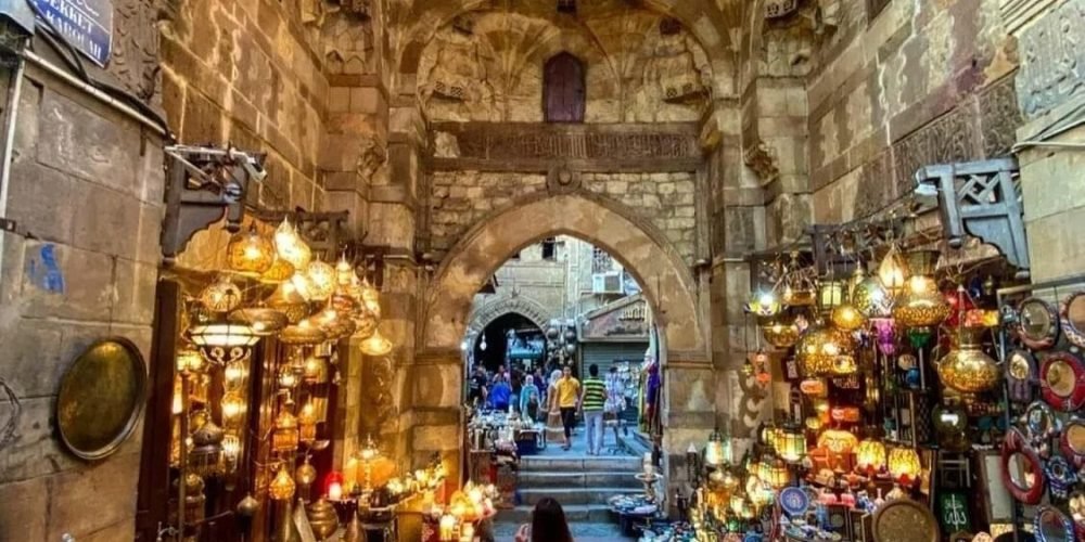A young woman in white walking through the historic Khan el-Khalili bazaar under ancient arched stone ceilings. The narrow alley is filled with glowing traditional lanterns, brass ornaments, and colorful souvenirs during an Egyptian Museum trip. Intricate Islamic architecture and vaulted stone entryways frame the bustling market scene in Old Cairo