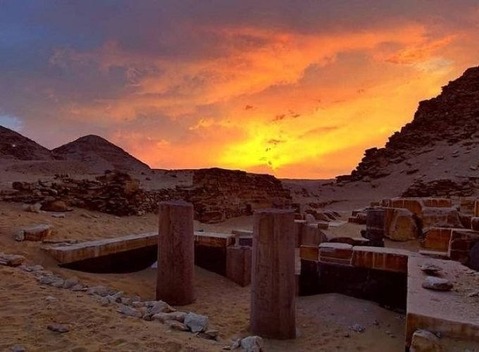 A breathtaking sunset view over the ancient ruins of Abusir. The foreground shows weathered stone pillars and excavation sites nestled in the desert sand. In the background, small hills rise against a dramatic sky filled with orange, yellow, and deep purple clouds as the sun sets on the horizon.