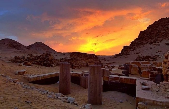 A breathtaking sunset view over the ancient ruins of Abusir. The foreground shows weathered stone pillars and excavation sites nestled in the desert sand. In the background, small hills rise against a dramatic sky filled with orange, yellow, and deep purple clouds as the sun sets on the horizon.