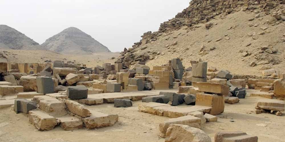 A wide, detailed view of the archaeological ruins at Abusir during the day. Scattered across the sandy desert floor are numerous large, weathered limestone and basalt blocks from ancient structures. In the background, a steep hillside covered in debris rises to the right, while two distant, triangular pyramid silhouettes stand against a pale, overcast sky.