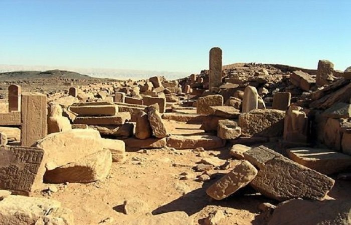A bright daylight view of the archaeological site at Abusir showing a dense field of scattered, weathered limestone blocks and ancient stone fragments. In the middle of the debris, a tall, slender stone stela or pillar stands upright. The background features a vast desert horizon under a clear blue sky with distant hills visible on the left