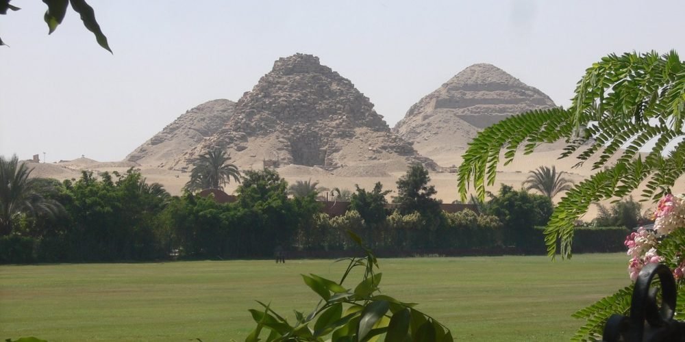 A scenic view of the pyramids of Abusir seen across a lush green lawn and garden. Two distinct, weathered pyramids rise from the sandy desert hills in the background under a bright, clear sky. In the foreground, green leafy branches and palm trees partially frame the shot, creating a peaceful contrast between the fertile land and the ancient desert monuments.