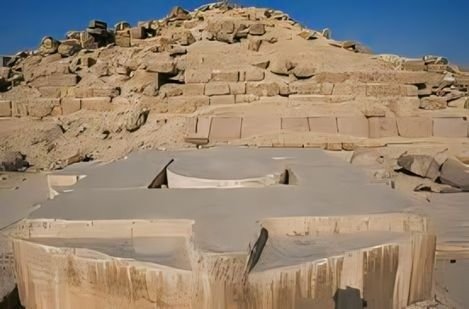 Au premier plan, une grande table d'offrandes circulaire et massive en albâtre poli gris clair, présentant des fissures naturelles et une cavité centrale. En arrière-plan, la pyramide à degrés de Djéser s'élève majestueusement, faite de blocs de calcaire ocre empilés, une vue saisissante lors d'une Excursion aux Pyramides