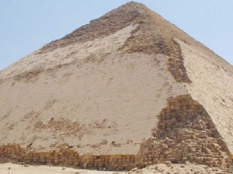 Une vue large à Dahshour montrant une immense pyramide de pierre aux faces triangulaires lisses et de couleur ocre. Le sommet est pointu et le ciel derrière est bleu très clair. Au pied du monument, sur le sable, on voit de petites silhouettes de personnes et un homme à dos de chameau lors d'une Excursion aux Pyramides