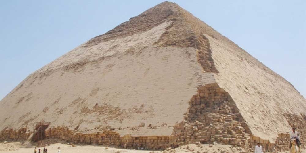 Une vue large à Dahshour montrant une immense pyramide de pierre aux faces triangulaires lisses et de couleur ocre. Le sommet est pointu et le ciel derrière est bleu très clair. Au pied du monument, sur le sable, on voit de petites silhouettes de personnes et un homme à dos de chameau lors d'une Excursion aux Pyramides