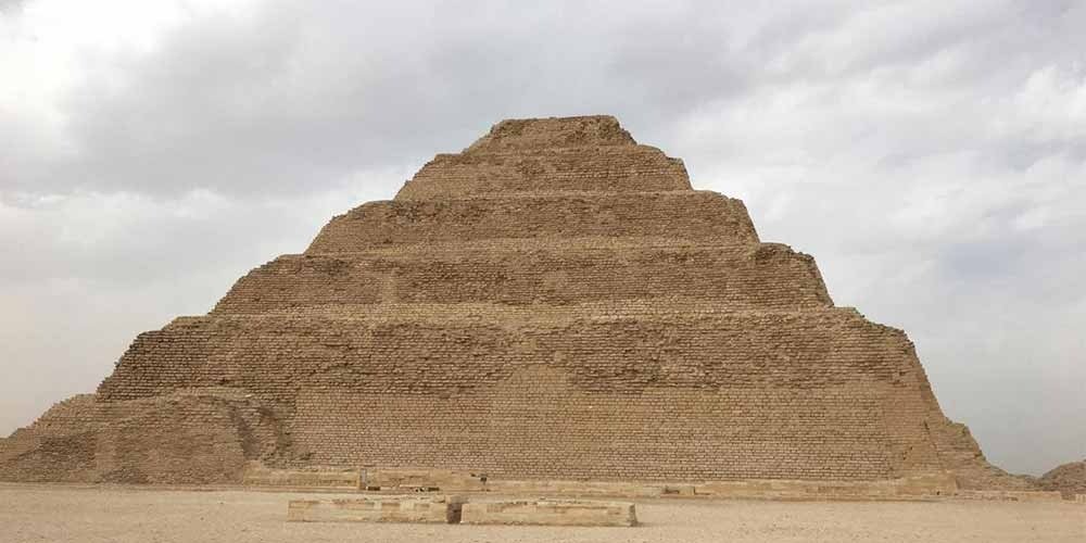 une vue de face d'une grande pyramide à six degrés construite en blocs de calcaire beige. La structure s'élève par paliers successifs au-dessus d'un sol de sable plat et clair, sous un ciel couvert de nuages gris, lors d'une Excursion aux Pyramides