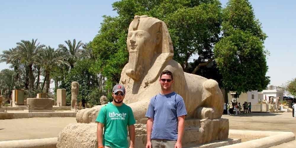À Memphis, deux jeunes hommes portant des t-shirts vert et bleu et des lunettes de soleil posent devant le grand Sphinx d'albâtre. La statue de pierre claire est sculptée avec un visage humain et un corps de lion, entourée de palmiers et d'arbres verts sous un ciel lumineux lors d'une Excursion aux Pyramides