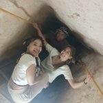 Trois touristes, deux femmes au premier plan et un homme derrière, sourient à la caméra tout en descendant ou montant dans un passage très étroit et incliné. Les murs sont en pierre brute de couleur beige et ils se tiennent à des barres métalliques jaunes fixées sur les côtés pour s'aider à avancer à l'intérieur des pyramides