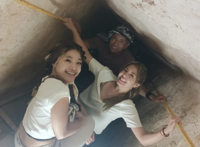 Trois touristes, deux femmes au premier plan et un homme derrière, sourient à la caméra tout en descendant ou montant dans un passage très étroit et incliné. Les murs sont en pierre brute de couleur beige et ils se tiennent à des barres métalliques jaunes fixées sur les côtés pour s'aider à avancer à l'intérieur des pyramides