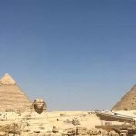 Vue panoramique en plein jour montrant le Grand Sphinx de Gizeh au premier plan، sculpté dans la roche calcaire. Derrière lui se dressent deux grandes pyramides de pierre massives sous un ciel bleu immense et dégagé. On aperçoit des vestiges de murs en pierre ancienne à gauche et une rampe de passage menant vers l'intérieur des pyramides
