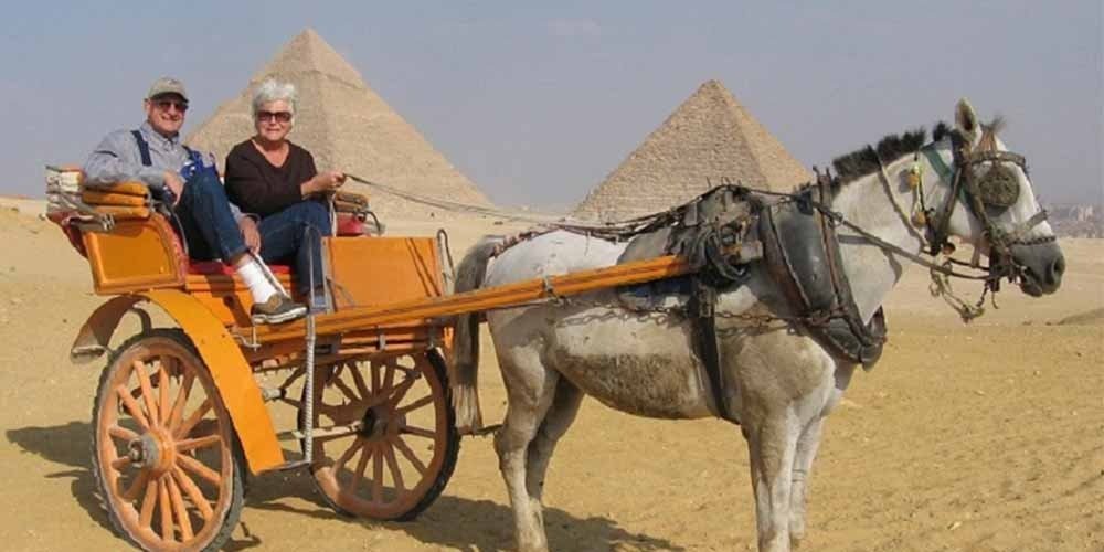 Un couple de touristes seniors، un homme et une femme، sont assis dans une calèche jaune et orange tirée par un cheval blanc. Ils se trouvent dans le désert de Gizeh، avec deux grandes pyramides s'élevant derrière eux sous un ciel voilé après une visite de l'intérieur des pyramides