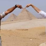 Un homme avec une chemise à motifs et une femme en chemise blanche et bandana posent dans le désert. Ils étendent chacun un bras vers l'autre pour que leurs index se rejoignent juste au-dessus du sommet de la grande pyramide en arrière-plan، créant un effet visuel amusant après avoir visité l'intérieur des pyramides