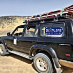 Une voiture tout-terrain 4x4 noire garée sur le sable doré d'un environnement désertique. Le véhicule est chargé sur son toit de tapis colorés et de matériel de camping attachés par des cordes vertes. On aperçoit des collines rocailleuses sombres en arrière-plan sous un ciel clair lors d'une expédition vers le désert blanc