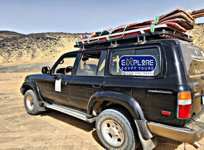 Une voiture tout-terrain 4x4 noire garée sur le sable doré d'un environnement désertique. Le véhicule est chargé sur son toit de tapis colorés et de matériel de camping attachés par des cordes vertes. On aperçoit des collines rocailleuses sombres en arrière-plan sous un ciel clair lors d'une expédition vers le désert blanc