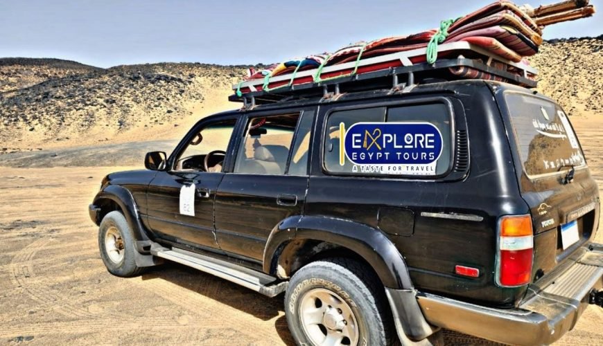 Une voiture tout-terrain 4x4 noire garée sur le sable doré d'un environnement désertique. Le véhicule est chargé sur son toit de tapis colorés et de matériel de camping attachés par des cordes vertes. On aperçoit des collines rocailleuses sombres en arrière-plan sous un ciel clair lors d'une expédition vers le désert blanc