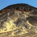 Une haute colline ou montagne de forme conique recouverte de roches volcaniques noires et sombres. Le sable jaune du désert remonte le long de ses flancs par endroits, créant un contraste de couleurs. Le sommet est escarpé et le ciel au-dessus est d'un bleu profond et sans nuages lors d'un voyage vers le désert blanc