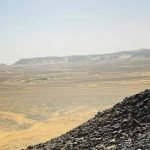 Une personne vue de dos se tient debout au sommet d'une colline escarpée parsemée de roches volcaniques noires. Elle contemple une vaste étendue désertique plate et sablonneuse qui s'étend jusqu'à l'horizon, où l'on aperçoit d'autres collines sombres et des plateaux rocheux sous un ciel blanc laiteux en route vers le désert blanc