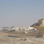 Une grande formation rocheuse de calcaire blanc pur en forme de champignon géant se dresse au milieu d'un paysage désertique plat. Le sol est parsemé de petits rochers blancs et de sable clair, avec d'autres structures rocheuses blanches visibles au loin sous un ciel bleu pâle et dégagé dans le désert blanc