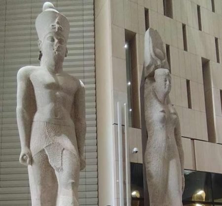 In the center of the hall stands the colossal 11-meter statue of Pharaoh Ramesses II, carved from red granite and standing on a modern stone pedestal. Behind the statue, a massive staircase with multiple levels of ancient Egyptian statues and artifacts rises toward the ceiling. The hall features contemporary architecture with tall, white geometric pillars and a high ceiling. Several tourists are seen walking around the base of the statue and ascending the stairs under the natural light coming from the museum’s glass façade, a highlight of the Experience Cairo for Group Day Trip
