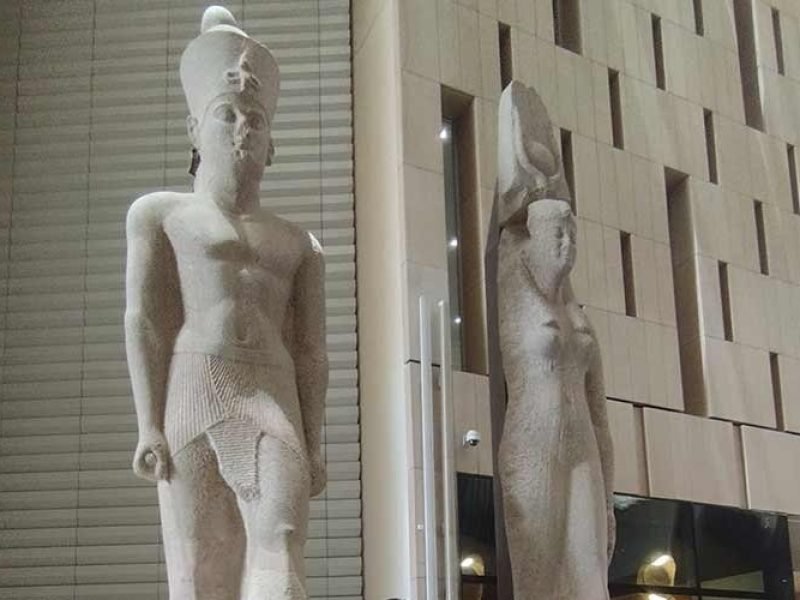 In the center of the hall stands the colossal 11-meter statue of Pharaoh Ramesses II, carved from red granite and standing on a modern stone pedestal. Behind the statue, a massive staircase with multiple levels of ancient Egyptian statues and artifacts rises toward the ceiling. The hall features contemporary architecture with tall, white geometric pillars and a high ceiling. Several tourists are seen walking around the base of the statue and ascending the stairs under the natural light coming from the museum’s glass façade, a highlight of the Experience Cairo for Group Day Trip