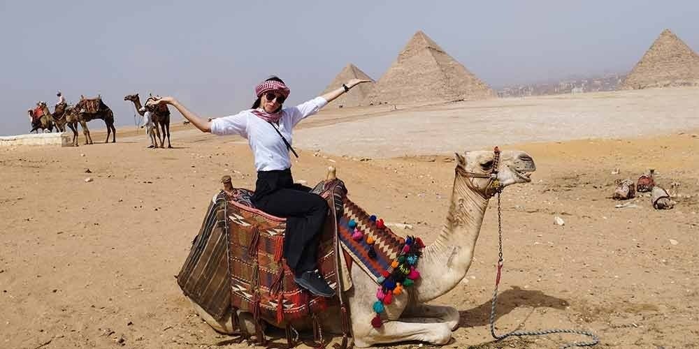 A female tourist sitting on a kneeling camel in the Giza desert, wearing a white shirt, black pants, and a traditional red-patterned headscarf. The camel has a colorful decorative saddle with pom-poms. In the sandy background, the three Great Pyramids of Giza stand under a clear sky, with other camels and tourists visible in the distance.