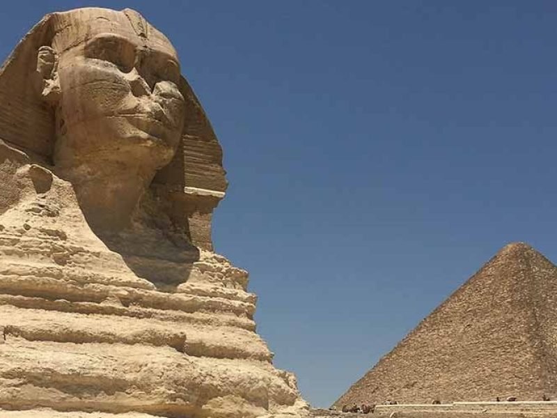 A close-up, side-profile view of the Great Sphinx showing its weathered limestone face and royal headdress against a clear blue sky. In the background, a large, majestic pyramid stands on the desert horizon under the bright Egyptian sun. The foreground features the tiered stone base of the monument, showcasing the ancient textures of the Giza plateau