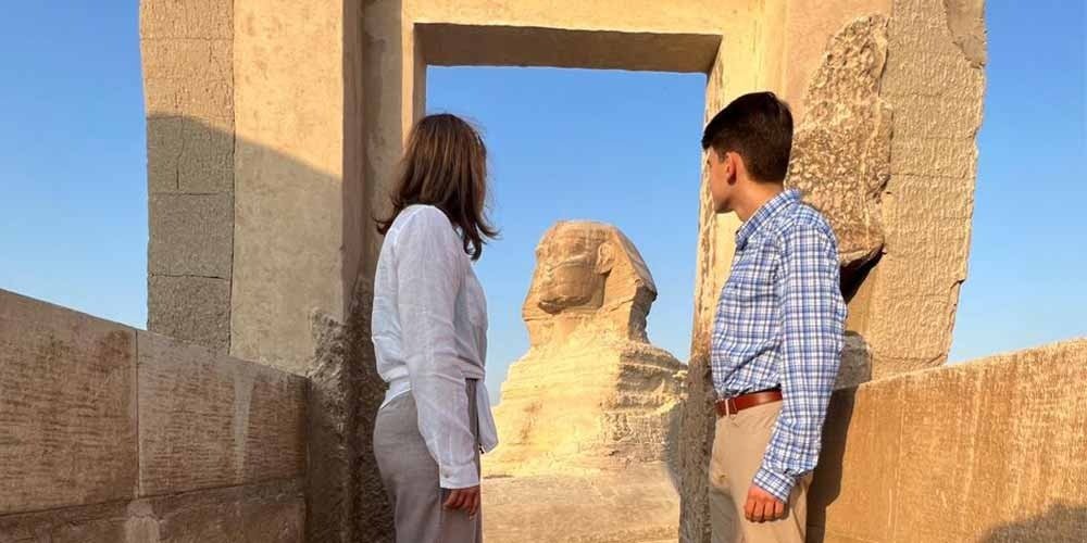 A young man in a blue plaid shirt and a woman in a white blouse stand between ancient stone pillars, looking out at the Sphinx through a rectangular doorway. The colossal limestone monument is perfectly framed in the center under a clear, warm sky during a private tour